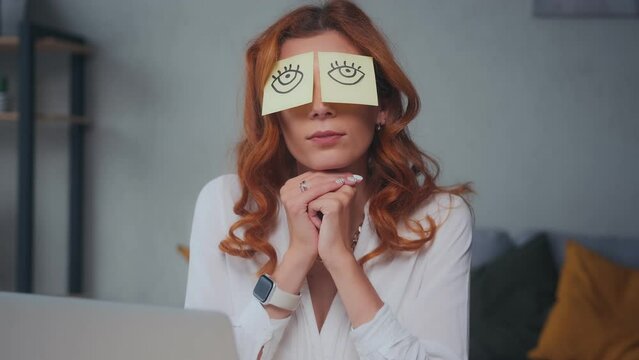 Young redhead Caucasian woman with funny stickers on eyes sleeps at workplace is lazy and shrinks from doing work sits on chair at office desk. Unprofessionalism, employee burnout