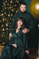 A stylish, smiling bearded man and a beautiful pregnant brunette girl sit and gently hug against the background of a decorated Christmas tree, balloons.