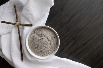 White bowl of ashes with white linen and small christian cross on a dark wood background with copy space