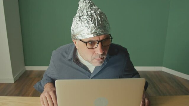 A Frightened Man in a Foil Cap Sitting at a Laptop, Anxiously Looking Around. Foil Hat For 5G Waves, Electromagnetic Fields, Mind Control, Mind Reading, Global Conspiracy. Mental Health.
