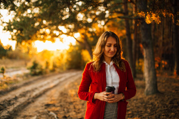 Elegant young girl at sunset in the forest. . Stylish girl in a red coat and skirt with coffee in her hands. Drinking coffee in nature. Coffee as a way of life
