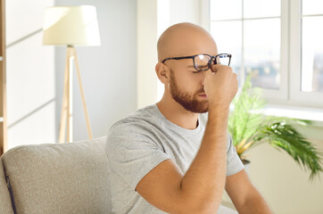Man who's sitting on sofa at home takes off his glasses and rubs his dry tired eyes. Young man...