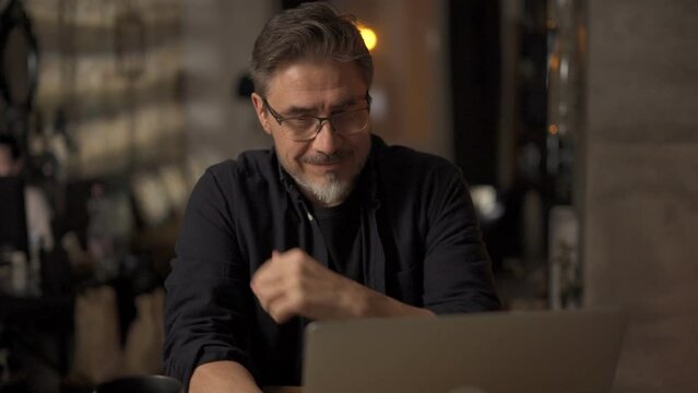 Happy mid adult man working with laptop computer in home office. Businessman smiling, thinking, wearing glasses.