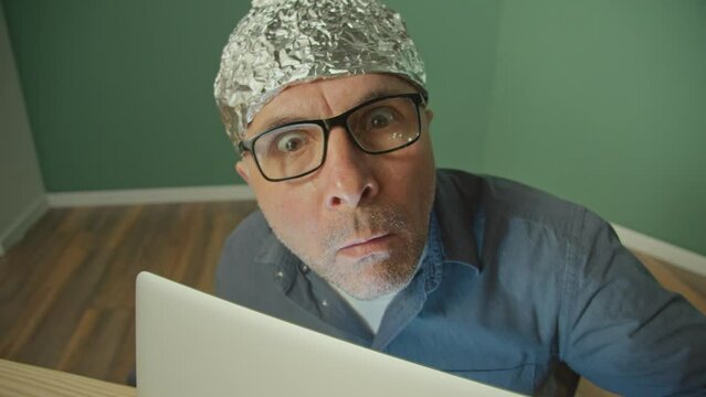 A Paranoid Man in a Protective Foil Cap Working on a Laptop Anxiously Peering Up. Fear, Espionage, Mind Control, Global Conspiracy. A Protective Helmet for the Brain From Aliens.