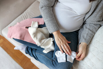 Top-down view of an unrecognizable Caucasian woman in third trimester of pregnancy sorting through baby clothing on a sofa