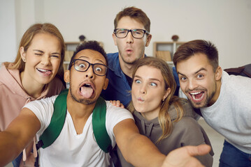 Happy students taking funny selfie indoors.Group of joyful young multiethnic friends having fun together, taking selfie on mobile phone, making hilarious grimaces, sticking tongues out, squinting eyes
