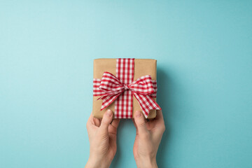 First person top view photo of saint valentine's day decorations female hands giving craft paper gift box with checkered ribbon bow on isolated pastel blue background with blank space
