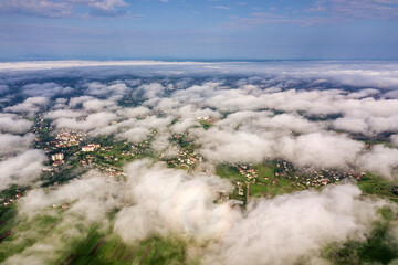 Aerial view of white clouds above a town or village with rows of buildings and curvy streets between green fields in summer. Countryside landscape from above.