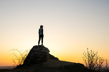 Silhouette of a woman hiker standing alone on big stone at sunset in mountains. Female tourist on high rock in evening nature. Tourism, traveling and healthy lifestyle concept.
