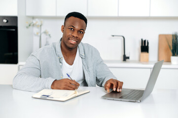 Attractive African American guy, student, freelancer, sits at a table in the kitchen, uses laptop and notepad for online learning, looks at the camera, listens to a lecture, takes notes,smile friendly