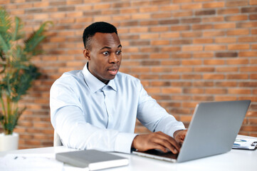 Shocked surprised African American business man, secretary, office worker, sitting at his desk in the office, looking at laptop in amazement, getting unexpected news, winning or making big profit.