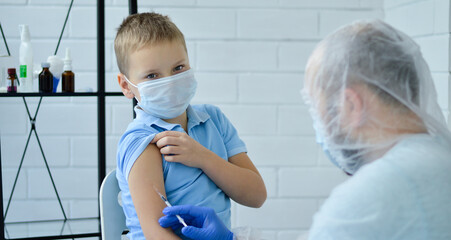 A doctor in blue gloves injects a vaccine with a syringe into a boy wearing a blue T-shirt in his...