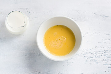 A white bowl with beaten egg and jug of milk on light blue background, top view. Cooking omelet,...