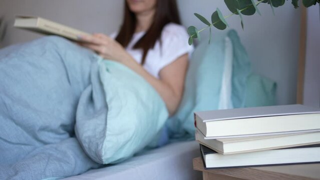 A pretty woman with dark hair sitting on the bed picks up a book to open and read enjoying a relaxing moment on the weekend. Concept mental health, healthy lifestyle