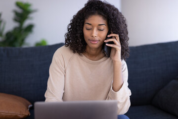 Young black woman at home talking on cellphone using laptop computer sitting on a couch