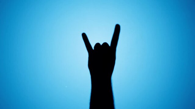 Man showing rock and roll gesture with fingers isolated on blue background. Male person making shadow silhouette with hand close-up.