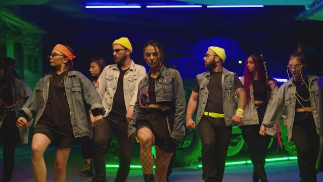 Group of stylish young people dancing synchronized dance  , moving near military tank and police car inside garage building Colorful stylish clothes  Modern professional dances . Green blue led lights