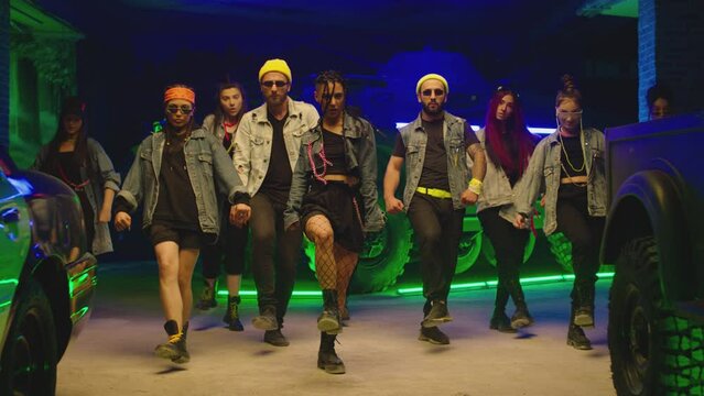 Group of stylish young people dancing synchronized dance  , moving near military tank and police car inside garage building Colorful stylish clothes  Modern professional dances . Green blue led lights