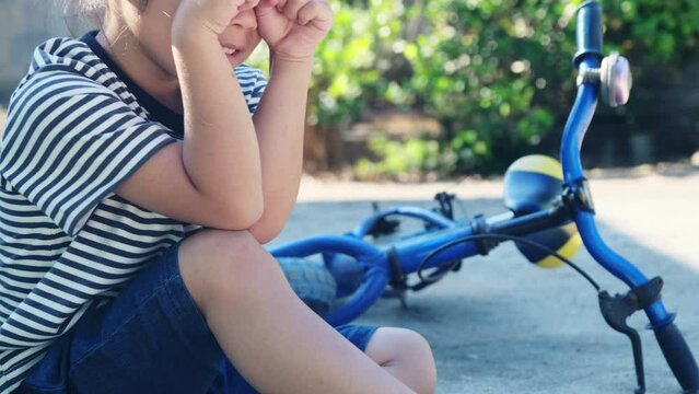 Sad Little Girl Sitting On The Ground After Falling Off Her Bike At Summer Park. Child Was Injured While Riding A Bicycle.