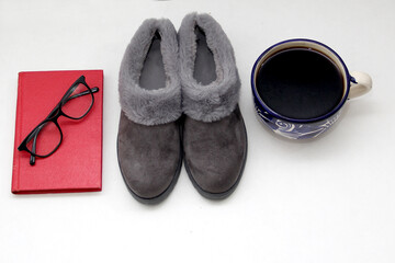 Gray fluffy, padded slippers, very comfortable next to a notebook, pen, book, glasses and coffee for a moment of relaxation and tranquility next to the bed
