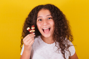 Afro brazilian, latin american girl with curly hair smiling, holding cheese and guava skewers, with...