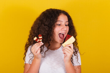 Afro-Brazilian, latin-american girl with curly hair smiling, holding skewers of cheese and guava,...