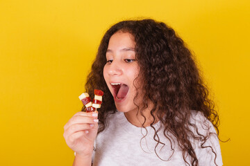 Afro brazilian, latin american girl with curly hair smiling, eating cheese and guava skewers,...