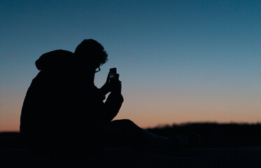 Silhouette of a Man sitting down at the side of the road during a fall evening taking a photo of the sunset
