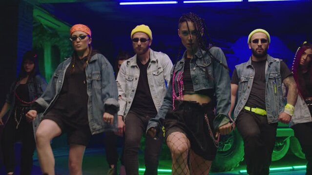 Group of stylish young people dancing synchronized dance  , moving near military tank and police car inside garage building Colorful stylish clothes  Modern professional dances . Green blue led lights