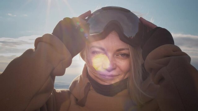 A beautiful girl puts on winter goggles or a mask for snowboarding. Big Blue eyes. Blond hair moving in the wind with slow motion. Getting ready for skiing on a snowy slope. Travel at the ski resort