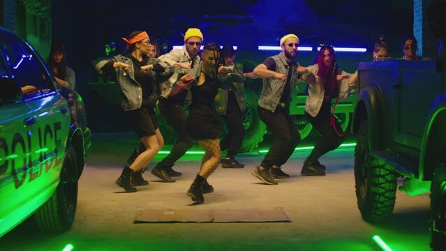 Group of stylish young people dancing synchronized dance  , moving near military tank and police car inside garage building Colorful stylish clothes  Modern professional dances . Green blue led lights