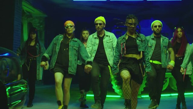 Group of stylish young people dancing synchronized dance  , moving near military tank and police car inside garage building Colorful stylish clothes  Modern professional dances . Green blue led lights