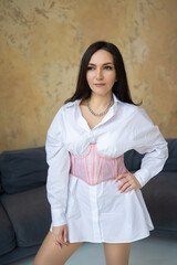 brunette girl in a white shirt-dress and a pink corsage sits on a gray sofa