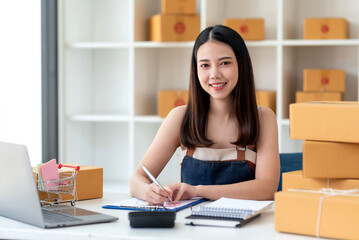 Portrait of young Asian small business owner working at home office writing address on cardboard box at workplace.  Smile woman is working at warehouse for online store looking at camera.