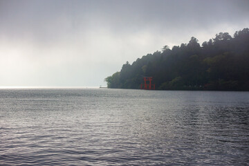Scenic dark view of Lake Ashi in Hakone, Japan with forest and red torii gate and cloudy sky background. No people.