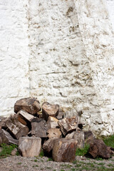 Chopped firewood lie in a pile at the base of an old stone wall. The white stone wall is lit by the summer sun. A pile of chopped firewood in on green grass.