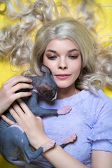 Portrait of young woman cosplay elf lying down on yellow background, gently clutching Sphinx kitten to her chest. Elf in blue dress, with beautiful blonde curly long hair, eyes of different colors.