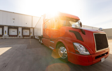 Wide angle of American Semi Truck parked at the dock. Tucking and logistics with red big rig waiting to get loaded at company. Shipping and receiving in the transportation business on a sunny day.