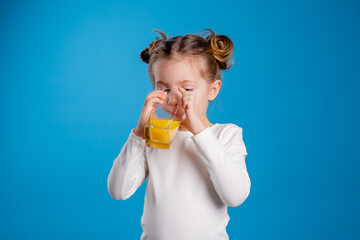 little girl with a funny hairstyle in a white T-shirt drinks juice from a glass. blue background. useful products for children. space for text. High quality photo