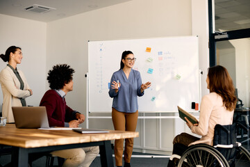 Young happy businesswoman gives presentation to group of diverse colleagues in meeting room.