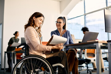 Happy businesswoman in wheelchair going through reports while working female coworker in the office.