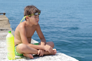 A cute boy in swimming glasses and shorts with a bottle of water sits on a pier on the seashore.