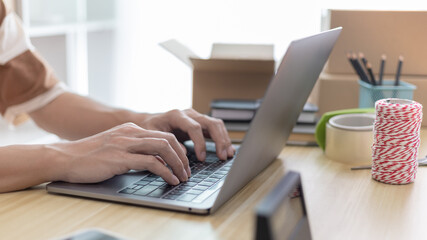 Young man uses a laptop to chat with customers who come to order product, Freelance work at home, Packaging on background, Sell online, Small business owner, Online shopping SME entrepreneur.