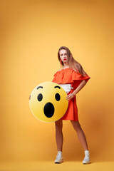 Social network concept. Pretty young woman holding yellow amazed emoji face. Colorful studio...
