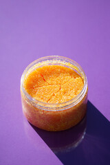 Open jar with orange sugar body scrub texture on a purple background. Skincare products , natural cosmetic. Beauty concept for body care