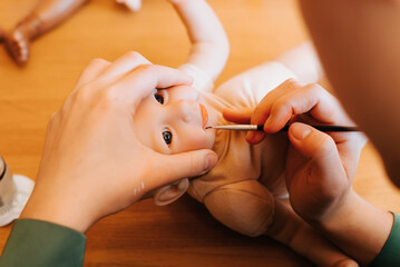 Reborning, puppet craft, hobby concept. Young female craftsman decorating realistic newborn doll sitting in workshop at home, painting lips on toy with brush, close-up. Selective focus on reborn face