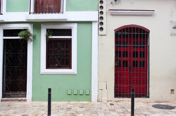 The entrance to the house with a red door leads to the entrance from the old stone-paved street. White and green house with doors and windows.