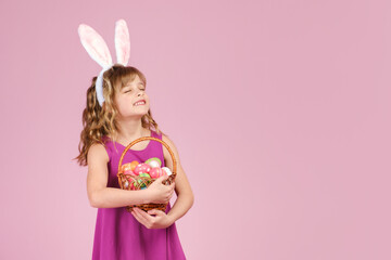 Little beautiful smiling girl in Easter bunny ears holds in hands a basket with eggs on pink background and raised his head with dreamy closed eyes. Copy space for text