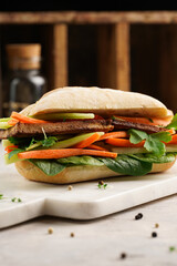 Traditional vietnamese banh mi sandwich with sliced smoked tofu, fresh spinach, shredded carrots and peeled cucumbers, cilantro on marble board on light grey textured background