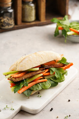 Traditional vietnamese banh mi sandwich with sliced smoked tofu, fresh spinach, shredded carrots and peeled cucumbers, cilantro on marble board on light grey textured background